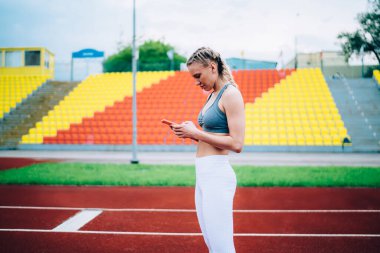 At kuyruklu ve ciddi yüzlü, yürüyüş pistinde duran ve sosyal medyada mesaj okuyan güzel, aktif bir kadının yan görüntüsü