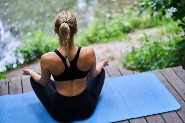 Mavi yoga minderinin üzerinde nilüfer çiçekli pozisyonda oturan ve nehir kıyısının bulanık manzarasına karşı ahşap terasta meditasyon yapan yüzü olmayan, zayıf, siyah aktif giysili bir kadın.