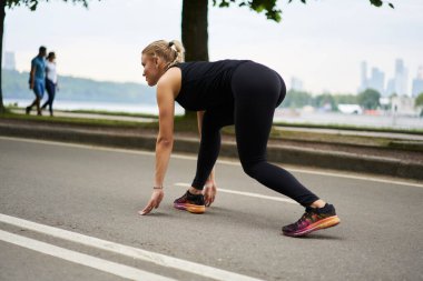 Düşük açılı, zayıf, kendine güvenen, siyah spor giyim ve renkli spor ayakkabılı bir kadın. Rıhtımda, deniz kenarında, bulanık şehir manzarasına karşı alçaktan koşmaya hazır.