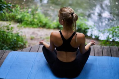 Arkadan bakıldığında, siyah spor kıyafetli, sakin, rahat bir kadın, Lotus pozu vermiş, Gyan Mudra 'yla mavi spor minderinin üzerinde oturuyor ve göl kıyısındaki ahşap terasta yoga yaparken gözlerini kaçırıyor.