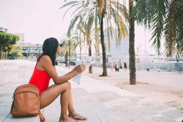 Side view of beautiful woman dressed in trendy outfit looking around after summer promenade in Spanish city with green palm trees, brunette female tourist with brown backpack resting with map