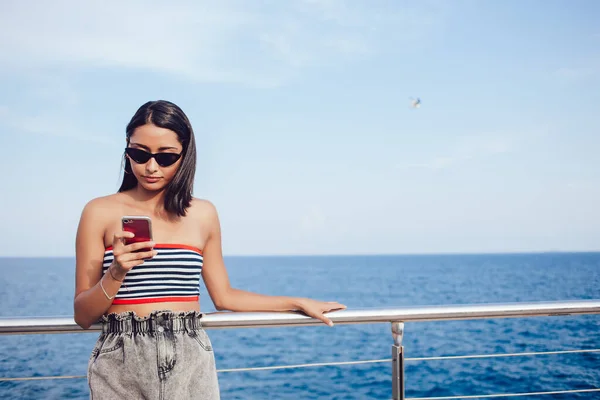 在公共网站上 身穿时髦太阳镜的千年女性利用现代蜂窝技术在海滨长廊在线浏览 年轻的拉美游客阅读旅游文章 — 图库照片