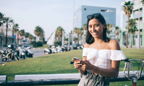Half length portrait of toothy Latino female tourist holding modern digital technology in hand and smiling during vacations journey to Spain, joyful millennial blogger enjoying smartphone using