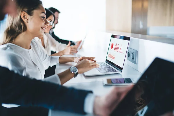 Side view of happy young lady cheerfully smiling and using laptop with various charts while sitting at table amidst coworkers in office