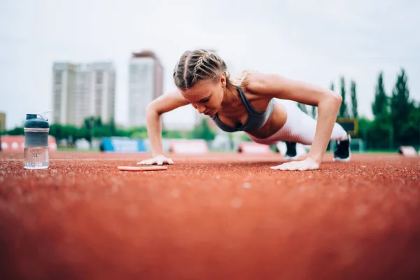 Pretty and purposeful woman full of motivation making effort and pushing up while training stamina at stadium on blurred background