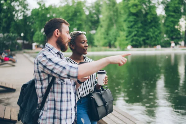 Side view of bearded guy pointing aside and talking with black girlfriend with beverage to go while standing near calm lake in green park