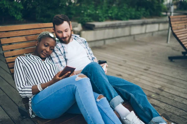 画面と笑顔に焦点を当てたカジュアルな服の中での高角度の大人の男アフリカ系アメリカ人のガールフレンドは公園の木製のベンチに座っているスマートフォンと対話しながら — ストック写真