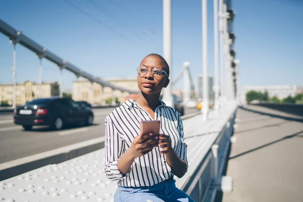 Stylowa Łysa Afroamerykanka Okularach Siedząca Telefonem Komórkowym Rękach Moście Miejskim — Zdjęcie stockowe