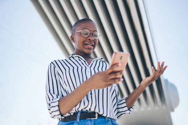 From below of surprised confused young African American female reading message on smartphone and spreading hands while standing under city bridge