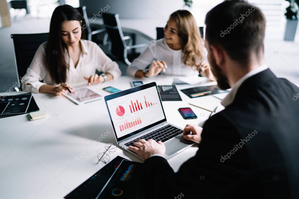 Blurred back view of male employee with beard browsing laptop with diagrams and statistics on screen sitting at table with women near smartphone
