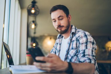 Kafede dizayn edilmiş iç dizaynı olan bir masada dizüstü bilgisayarla çalışırken gömlekli, sakallı ciddi bir adamın cep telefonuyla eğlenmesi. 