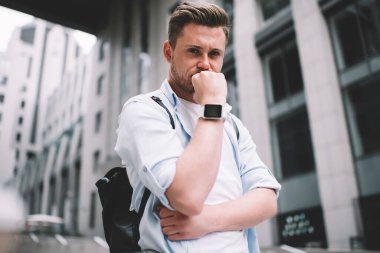 Genç, sakallı, akıllı, beyaz tişörtlü, mavi gömlekli, beyaz saçlı, şehirde dikilip kameraya bakarken çenesine yumruk atıp düşünen bir erkek.