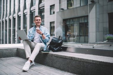 Genç, kablosuz kulaklıklar takan, dizüstü bilgisayar kullanan, kahve içen ve caddedeki taş bankta oturup gözlerini kaçıran neşeli bir adam.