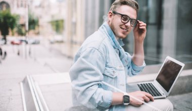 Gündelik giyinmiş, akıllı saat takan, dizüstü bilgisayar kullanan, sokakta oturup kameraya bakan parlak erkek camına bak.