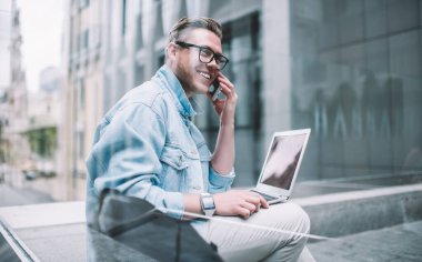 Gündelik giyinmiş, dizüstü bilgisayarlı, akıllı telefon kullanan ve şehir sokaklarında otururken iş arkadaşlarıyla konuşan gülümseyen genç bir adamın camından bakın.