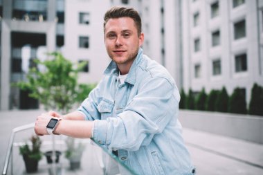 Yakışıklı, kendine güvenen, sıradan giyinmiş, akıllı saati olan şehir sokaklarında cam çitlere yaslanmış kameraya bakan genç bir adam.