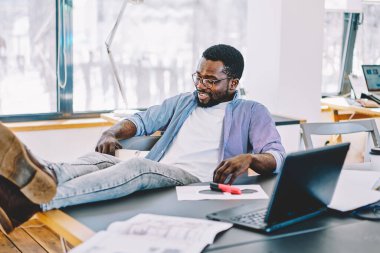 Rahat siyah adam gündelik kıyafetler ve gözlüklerle gülümsüyor ve modern işyerinde dinlenirken bacakları masanın üstünde tutuyor.
