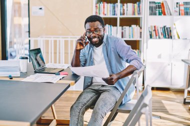 Gülümseyen, beyaz dişli, sıradan giyinmiş, cep telefonuyla arayan, elinde belge tutan, Afro-Amerikan bir erkek. 