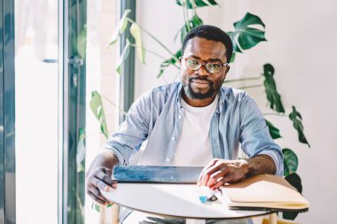 Güneşli odada kameraya bakarak akademik projeye hazırlanırken tablet üzerinde çalışan düşünceli, düşünceli siyahi adam.