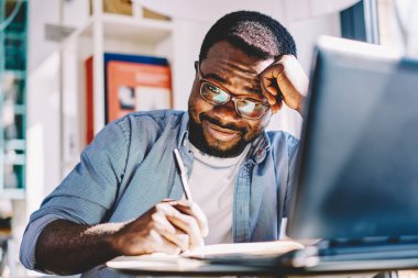 Afrika kökenli, yorgun, neşeli, çalışkan bir adam ders için makale yazarken iş yerindeki laptopun önünde kafa üstü yatıyordu.
