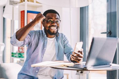 Gözlüklü, sıradan giyimli, neşeli Afro-Amerikan erkek. Zaferi kutlarken dizüstü bilgisayar ve kağıtlarla masada otururken evde akıllı telefon kullanıyor.