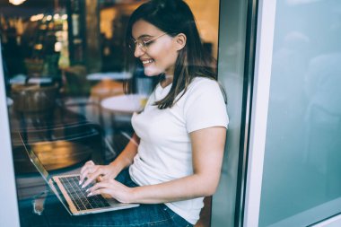 Üniversite kafesinin penceresinin yanında otururken bilgisayarında daktilo yazan gözlüklü neşeli genç kız öğrenci camından yan bakış.