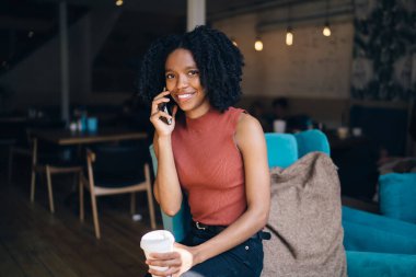 Neşeli kıvırcık saçlı, günlük giysiler içinde, cep telefonuyla konuşan, yazın açık terasta, mavi kanepede bir fincan kahve içen bir etnik kadın.