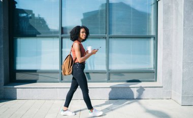Sırt çantasındaki kahve fincanıyla şehirde yaz aylarında sokakta cep telefonundan mesajlaşan sıska, etnik bir kadın.