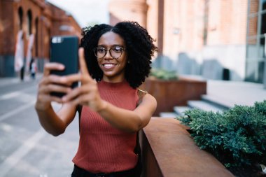 Afro saçlı, sıradan giyinmiş, gözlüklü, cep telefonu kullanan ünlü şehrin caddesinde selfie çeken mutlu bir zenci kadın.