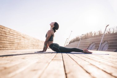 Kobra yoga pozisyonunda rahat spor giyimli huzurlu bir kadın parlak güneş ışığında minder üzerinde pratik yapıyor.