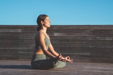 Nilüfer çiçeği pozisyonunda sakin bir kadın, dizlerinin üzerinde elleri ve gün doğumunda ahşap patikada gözleri kapalı.