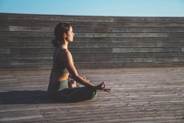 Nilüfer pozisyonunda oturan, dizlerinin üzerinde el ele tutuşan ve sabah güneşinde ağaçtan terasta meditasyon yapan rahat etnik kadının yan görüntüsü.