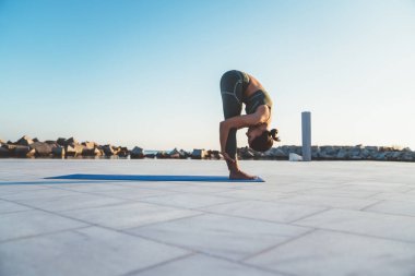 Yeşil spor giyim giymiş esnek kadın yoga yapıyor ve sabah mavi paspasın üzerinde öne eğilip poz veriyor.