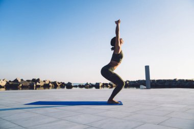 Spor elbiseli genç bir kadının spor kıyafetleri içinde spor minderinin üzerinde durup arka planda sandalye yogası pozu vermesi.