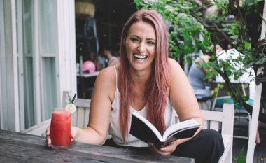 Neşeli gülen kadın kitap okuyor ve yaz günü açık terasta masada otururken taze soğuk smoothie içiyor.
