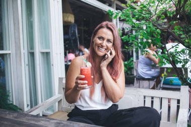 Güzel, uzun saçlı, beyaz tişörtlü, elinde meyve püresi olan bir kadın. Sıcak yaz gününde kahve terasında oturmuş konuşuyor ve telefonda gülüyor.