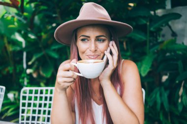 Neşeli gülümseyen, şapkalı, akıllı telefonlu ve kahvesini yudumlayan modern kadın tropikal yaz gününde açık terasta oturuyor.