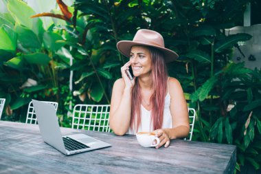 Yazın açık terasta bir fincan kahveyle masada otururken şapkalı, akıllı telefonlu ve bilgisayarlı konuşan mutlu bir kadın.