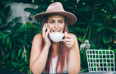 Gülen modern kadın, tropikal yaz gününde açık terasta otururken akıllı telefondan konuşan ve kahve içen şapkalı.