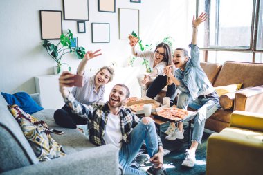Dost canlısı gençler pizza yiyor ve cep telefonlarında selfie çekiyor. El kol hareketleri yapıyor ve iki parmağını kanepe ve yerde bacak bacak üstüne atmış bir şekilde gösteriyor. 