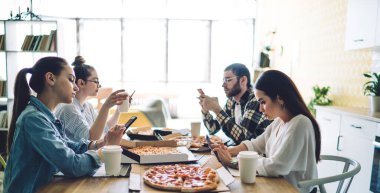Ciddi arkadaşların toplantı sırasında cep telefonlarını karıştırıp matarada taze pizza ve kahveyle masaya oturmalarının yan görüntüsü.