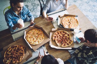 Yukardan taze pişmiş iştah açıcı pizzalar. Yemek masasında oturan ve cep telefonlarını karıştıran mahsul aç arkadaşlar için. 