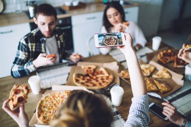 Yüksek açılı seçici bir odak noktası var. İsimsiz bir kadın, pizza çekerken arkadaşlarıyla masada oturuyor ve parti sırasında yemek yiyor.