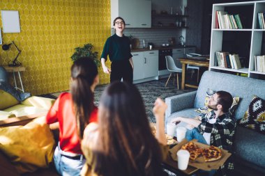 Gündelik arkadaşlar iştah açıcı pizzalar giyer ve modern apartmandaki yumuşak rahat koltukta dinlenirken gülen bir arkadaşa bakarlar.