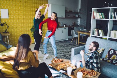Güneşli odada pizza ve tek kullanımlık fincanlar dağıtırken ayakta durup el kol hareketi yapan kadınları eğlendirirdi.