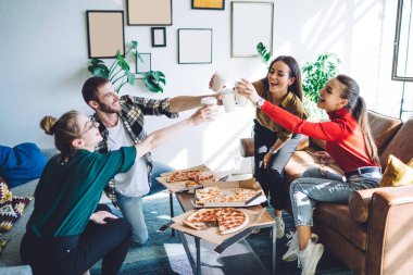 Modayı takip eden neşeli iş arkadaşları ofiste parti yaparken pizza yiyip kağıt bardakları tokuşturuyorlar.
