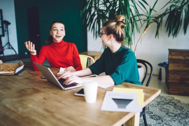 İyimser genç bir bayan masada oturmuş, ev ödevlerini yaparken arkadaşlarıyla konuşurken gülümsüyor ve el kol hareketleri yapıyor.