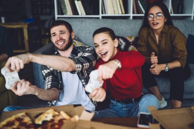 Sıradan giyinen konsantre arkadaşlar heyecan verici video oyunları oynarken rahat kanepenin yanında takılıp modern dairede pizza yiyorlar.