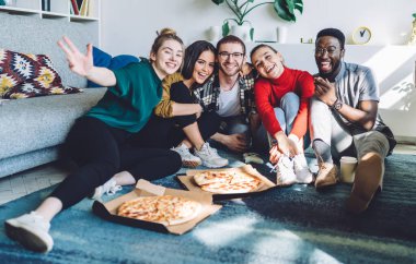 Neşeli çoklu etnik arkadaşlar, günlük kıyafetler içinde pizza ve paket servis içkileriyle yerde oturup gündüz partisinde kameraya bakıyorlar.