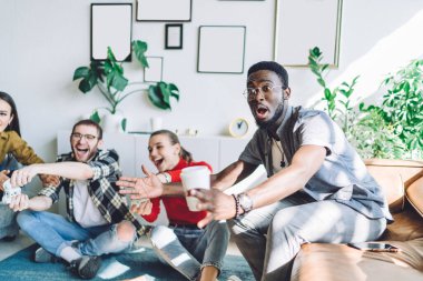 Gözlüklü ve günlük giysileri olan ağzı açık Afro-Amerikan bir adam ev partisinde oyun oynayan arkadaşları hakkında endişeleniyor ve onları izliyor.